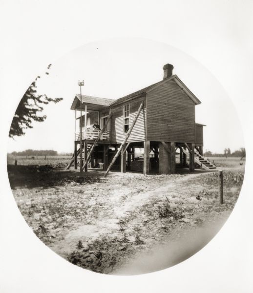 Exterior view of an Illinois farmhouse on stilts above the Saline River. A woman poses on the porch. Wood beams are shoring up the sides of the house, and a birdhouse is perched on top of a wood post.