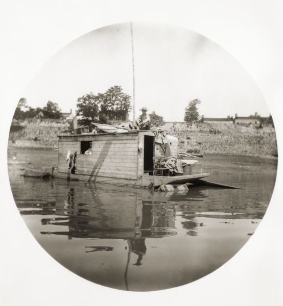 A man is seated on the roof of his shanty boat. Another person is at a window. Shoreline with buildings in background.