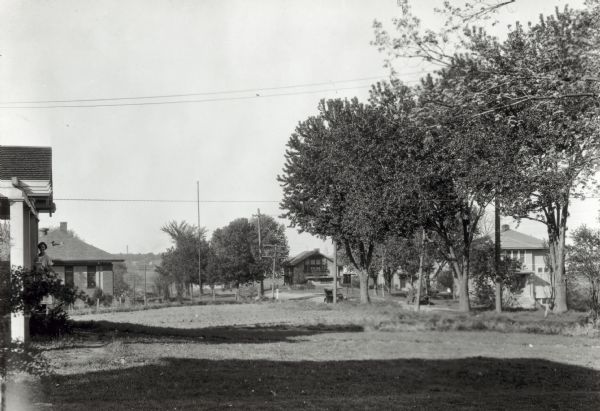 View of Nakoma Road looking northeast, with several houses and trees, and automobiles parked on the street. There is a woman standing on the steps of a porch on the left.