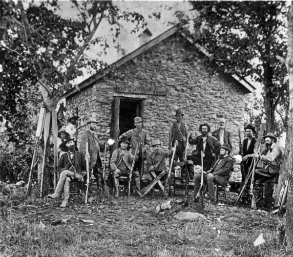 Group of men posed in front of small stone building with guns. Sign above doorway says "Waupun Hunters Club Home."