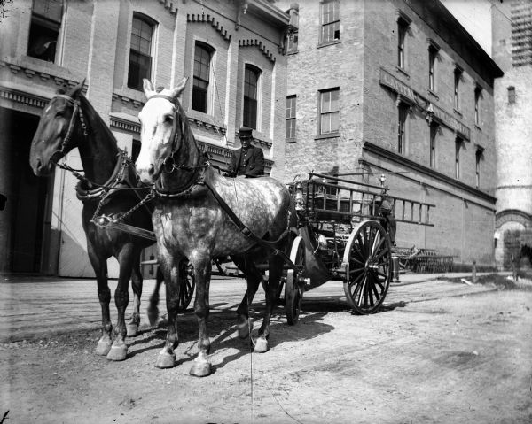 Horse-drawn fire wagon in front of several brick buildings, including the Capitol Publishing building, near the water tower. The man driving the wagon may be a firefighter.