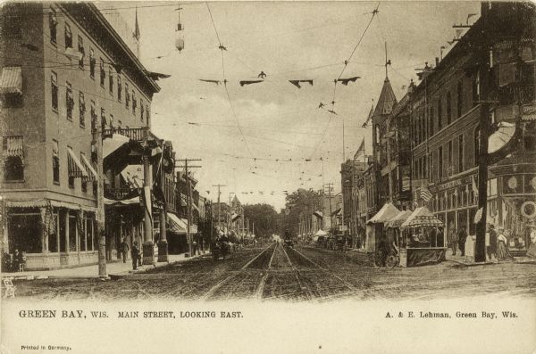 Main Street, looking east at storefronts. Booths with flags and bunting are set up on the sidewalk. Caption reads: "Green Bay, Wis. Main Street, Looking East."