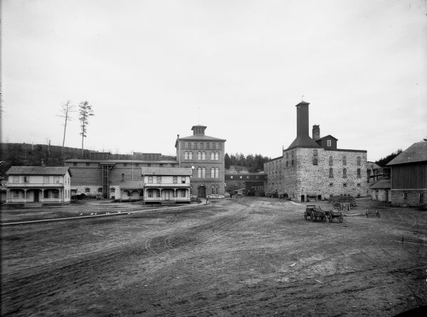 Buildings and grounds of the Leinenkugel Brewery. There are beer wagons in the yard, chickens along a wooden walkway, and men on a horse-drawn wagon near a loading dock.