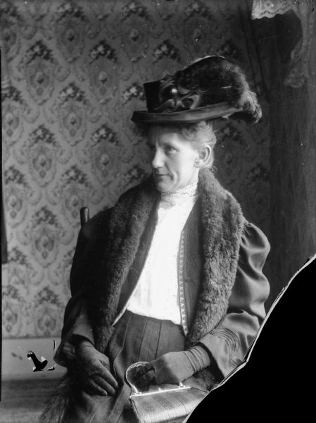 A woman seated in a chair wearing a coat with a fur collar, hat, and holding a handbag.