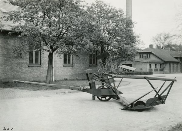 Left side of McCormick reaper of 1849, sitting in a street, with two buildings, a smokestack, and trees behind.