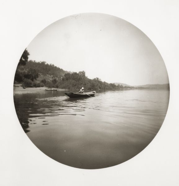 A woman rowing in a boat on the Ohio River. There is another boat on the shoreline behind her.