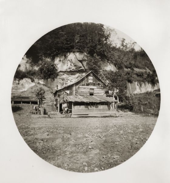 Exterior view of a log house at the base of a cliff. A man and a boy pose in front of the house.