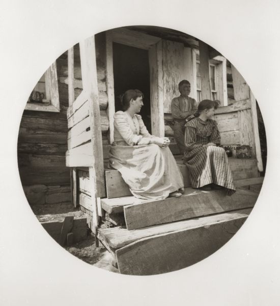 A father and his two daughters are seated on the porch of their log house.