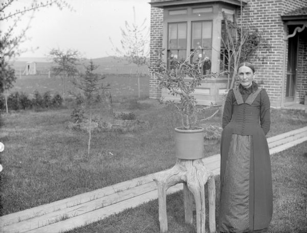 Outdoor portrait of a woman in a long dress posing with a crown of thorns plant in the front yard of house. The plant is in a pot which is sitting on a piece of driftwood near a board sidewalk.