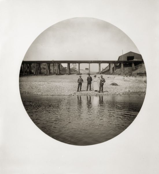 View from the Ohio River of four friends (three men and one woman) standing on the shore, seeing the travelers off. There is a bridge in the background.