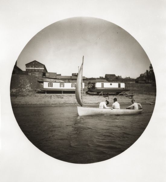 View from river of three men in a sailboat near the shoreline. Behind them are houseboats up on blocks on the shore of the Ohio River, with other buildings and a railroad car on the hill behind them.