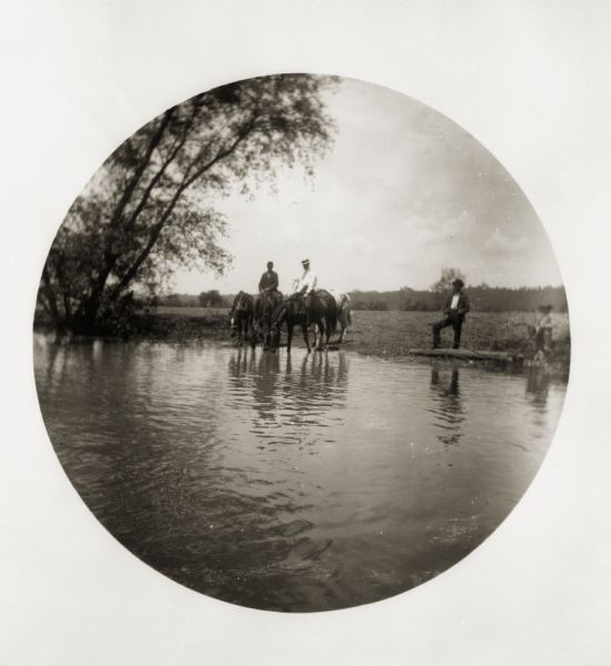 Farm boys watering horses. Two of the young men are seated on horseback while a third stands near them.