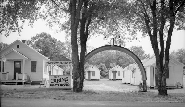 Entrance sign and rainbow between two trees in front of several cabins at the Rainbow Cabins.  The cabins were located on Route 16 on Vine Street.
