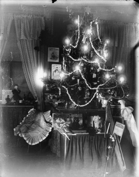 A Christmas tree on a tabletop is lit up in a dark living room, possibly in the home of Frederick King Conover. Toys, including a top, sled, and books, are displayed on the table and floor.