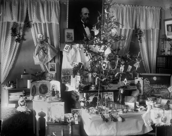 A decorated Christmas tree sits on a tabletop, possibly in the home of Frederick King Conover. Toys, including dolls, books, miniature furniture, cover tables in front of the tree.