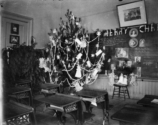 Interior view, with desks in the foreground, of an elementary classroom decorated with a Christmas tree near the blackboard, and other holiday decorations, including dolls, portraits, and signs.