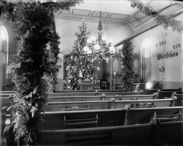 Interior view of the Old Moravian Church at 6th and Cole Streets decorated for Christmas. There is a very tall Christmas tree behind the pulpit, and other greens on posts and swags among the pews. Part of an organ can be seen on the right near the front of the church, and an ornate chandelier hangs from the ceiling.