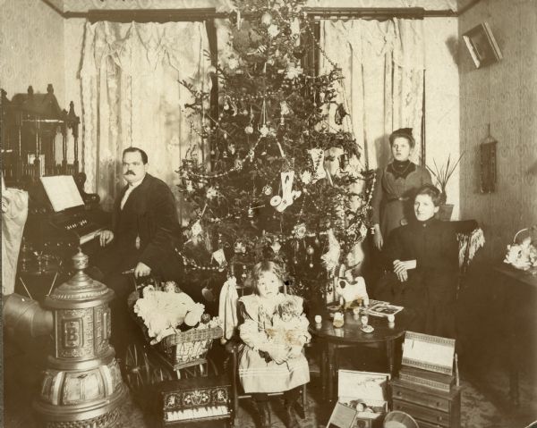 A formally dressed family pose around a Christmas tree, probably in their living room. A man is sitting at a piano on the left side, with his hand resting on a baby carriage full of dolls, a young girl sits in a chair with a doll, and two women are posing on the right.
