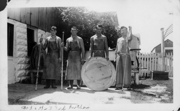 Casper Jaggi, on right, with cheese harp, and three brothers of the Erb family, (holding stirrers), standing outdoors at the Coldren Cheese factory. They are all wearing long aprons and boots.