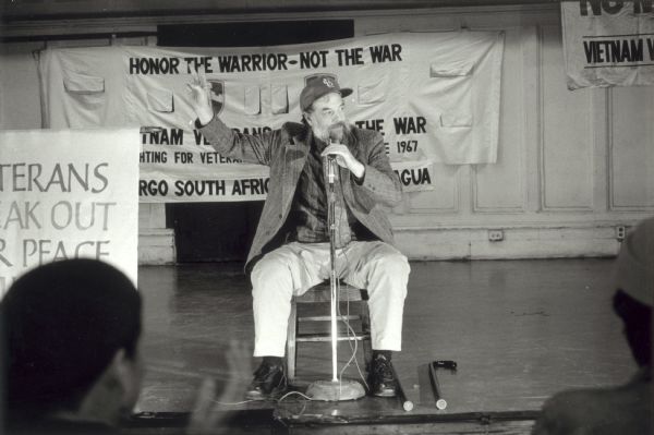 Brian Willson, Vietnam veteran who lost both legs during a demonstration in 1987, holds up his arm to give the peace sign to audience at a New York peace conference in 1988.  The Vietnam Veterans Against the War banner behind him indicates the VVAW's ongoing opposition to American foreign policy. Willson was not an anti-war activist during the Vietnam era, but he gradually became one in the peace movement and lost his legs as a result of a demonstration against U.S. actions in Nicaragua.