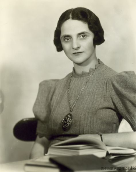 Portrait of Toni Sender seated with books. She is wearing a necklace with a large pendant.