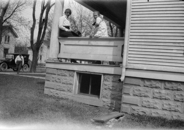 A woman and a child sit on a railing and look out from the porch of their house. A man is in the background approaching from a car parked in the street.