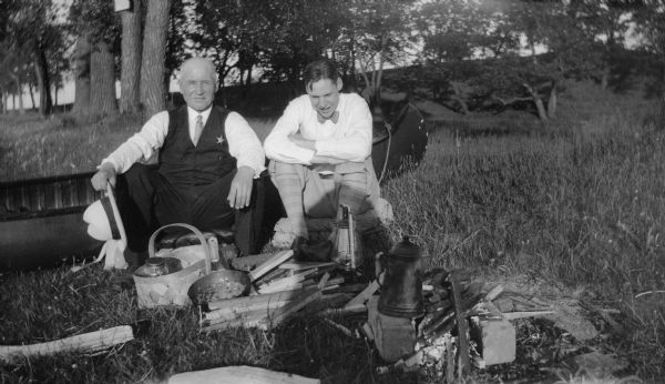 Edward J. Rickers with UW nightwatchman "Gramps," on a Lake Mendota outing. They are seated on the grass together with a picnic lunch and a campfire. There is a canoe behind them.