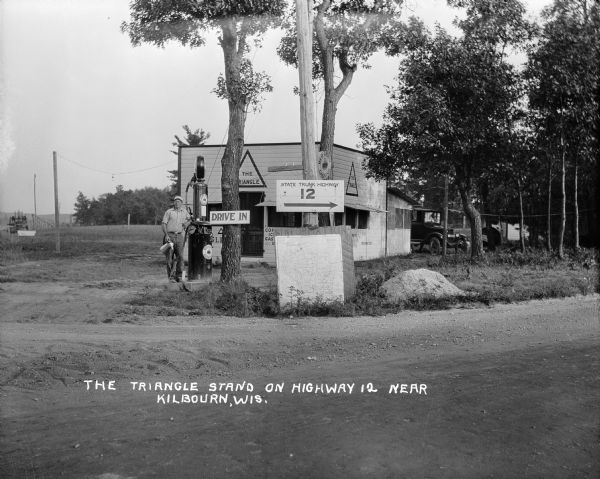 Exterior view of the Triangle filling station on Highway 12, which sold gasoline, ice, and other items. A man stands in front by the gas pump, and a map of Wisconsin on a board is displayed under the sign for "State Trunk Highway 12."