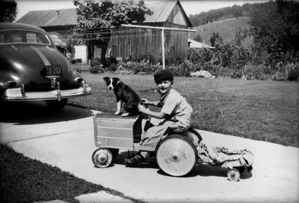 A young boy and his little dog sit together on a toy tractor in a driveway behind a parked car.