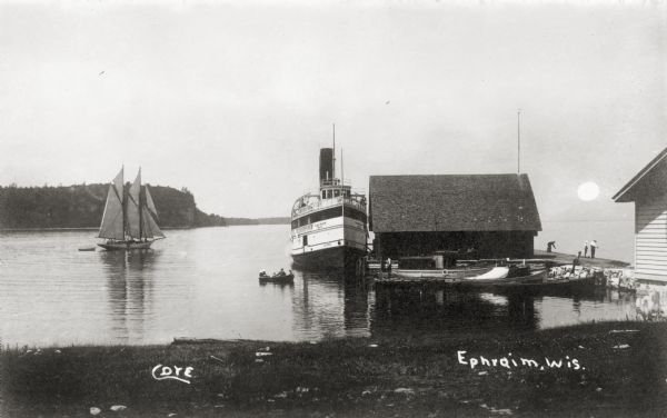 View from shoreline of boats docked at the fishing wharf. People are fishing from the pier, and a group of people are in a rowboat near a ferry. There are people a sailboat with a skiff in the water on the left. The bluff on the horizon is in Peninsula State Park Caption reads: "Cove, Ephraim, Wis."