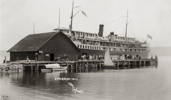 View across water towards a large ferry docked at the fishing wharf. A large group of people are standing on the pier, and passengers are standing at the rails of the ferry. The bluff on the horizon is in Peninsula State Park. A sailboat and other small watercraft are docked on the near side of the pier, and an automobile is parked near a building. Caption reads: "Ephraim-Wis."