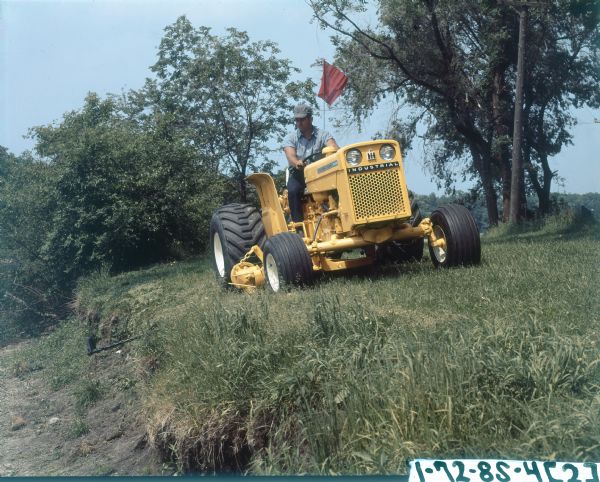 View of a man riding an International 2444 tractor with a No. 110 Balanced Head Mower. The tractor is mowing along the edge of bank.