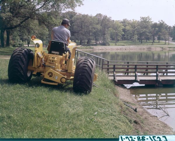 Color photograph of a man driving an International 2444 tractor with a No. 110 Balanced Head Mower.  The tractor is mowing near the shoreline of a pond or river.