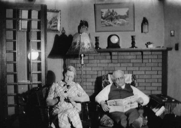 E.W. and Mary Brandel relax together in their home in front of a brick fireplace. They are both wearing eyeglasses; he is reading a newspaper and she is knitting.