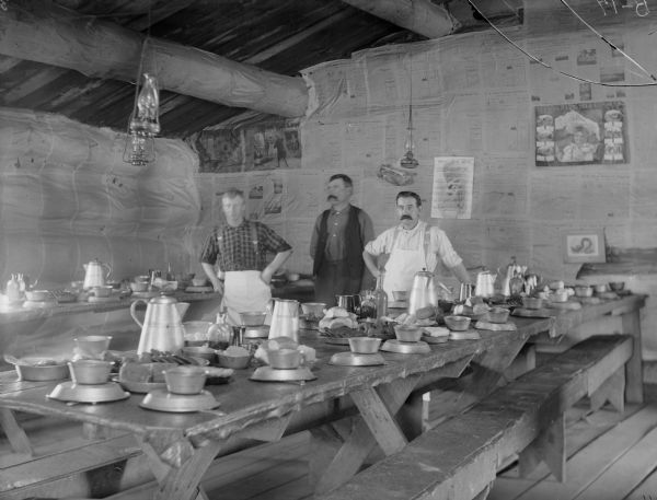 Interior of cook shanty, with oil lamps hanging from the ceiling beams. The back wall is covered with newspapers. Three men stand behind wood benches and tables laid with metal dishes.