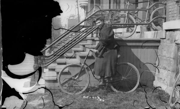 Woman sitting on a bicycle in front of a stoop with metalwork railing. Another bicycle sits on the porch behind her. She is wearing a hat, and a long skirt with a puffy-sleeved jacket.