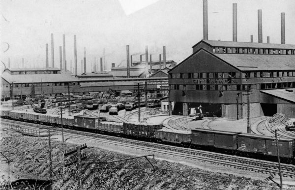 Elevated view of industrial buildings identified as smelting furnaces. A railroad train and railroad track runs the length of the foreground of the image and several lead into a railroad yard near the buildings.
