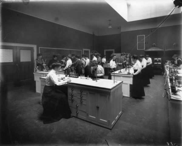 University of Wisconsin-Madison Home Economics Department Class. The women are using chemistry equipment and microscopes. On the blackboard is a formula for wood staining mixture, and there is a skylight in the ceiling.