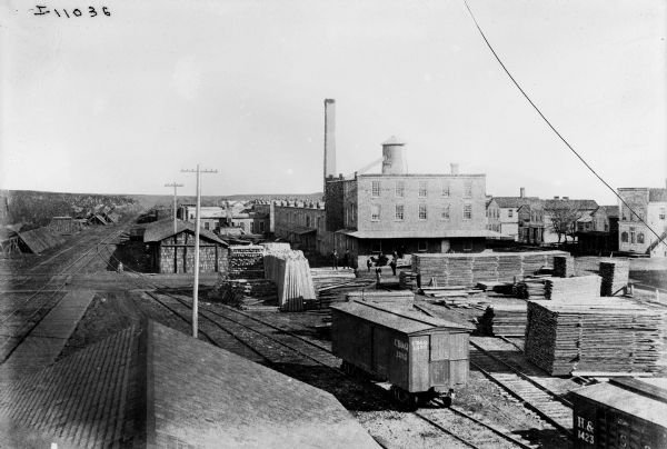Elevated view of men standing around a lumber yard among buildings at an International Harvester factory(?) site, most likely in Illinois. A Chicago, Burlington and Quincy railroad car is visible in the foreground. Railroad tracks run to the horizon on the left.