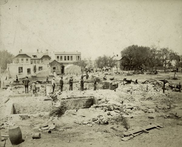 People stand amidst the ruins of Middleton after a fire, which burned 19 buildings. Debris litters the ground and foundations of buildings are exposed. There are some intact buildings in the background.