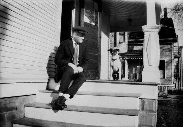 A man in a hat sits on the steps of the back porch of the Brandel family house with the family dog, Max.