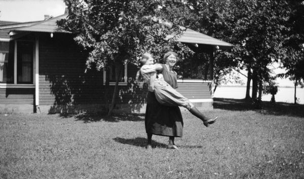 A woman playfully spins another woman (probably her daughter) in a yard in front of a house. There is a lake in the background.