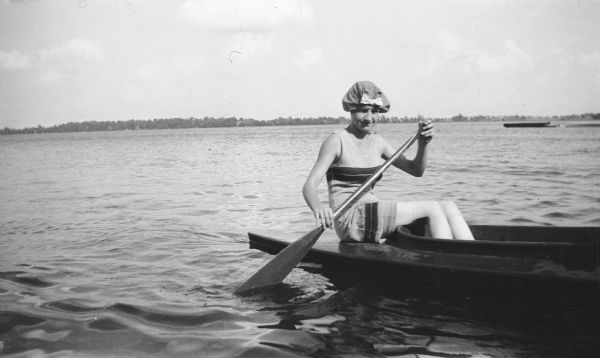 Mary Brandel paddles a canoe (or kayak) on Fox Lake. She is wearing a bathing suit and hat.