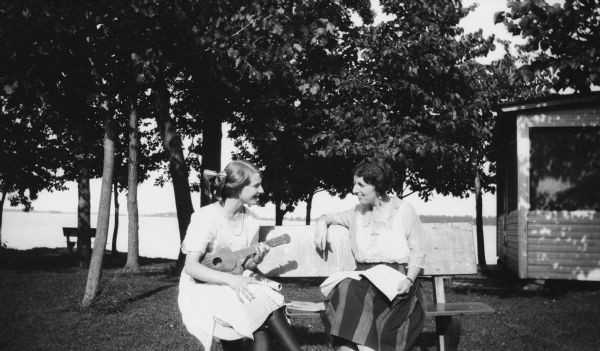 Mary Brandel sits at a picnic table with another woman. Mary is holding a ukulele and there is a cottage and lake in the background.