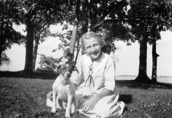 Mary Brandel sits outdoors on the grass with her dog Max. There is a lake in the background.