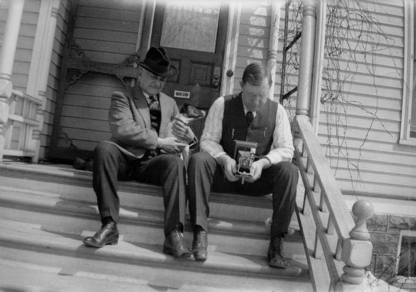 E.W. Brandel sits on porch steps holding his daughter's dog Max, a fox terrier. Next to him, holding a camera, is Dr. A.W. Jones, the twin brother of the photographer.