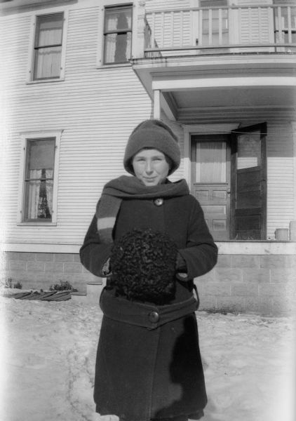 Ethel Walker, friend of Mary Brandel, poses in the backyard with a  house in the background. She is dressed for cold weather and wears a hat, scarf, and coat and has her hands in a muff.