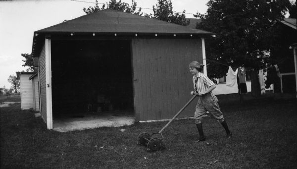 A fashionably dressed young woman mows the lawn with a push mower at the cottage. She is in front of an open shed, and a clothesline hangs from it. A house is in the background.