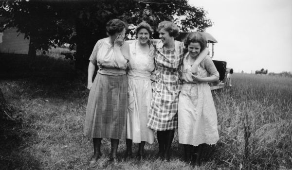 Four women pose together outdoors. There is an automobile behind them.