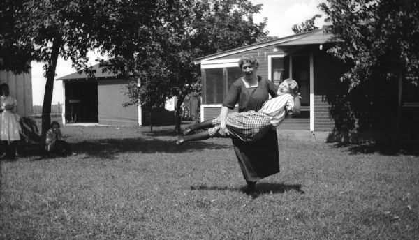 A woman holds a young woman (Mary Brandel?) in her arms in front of their cottage at Fox Lake. Two other people can be seen on the left.